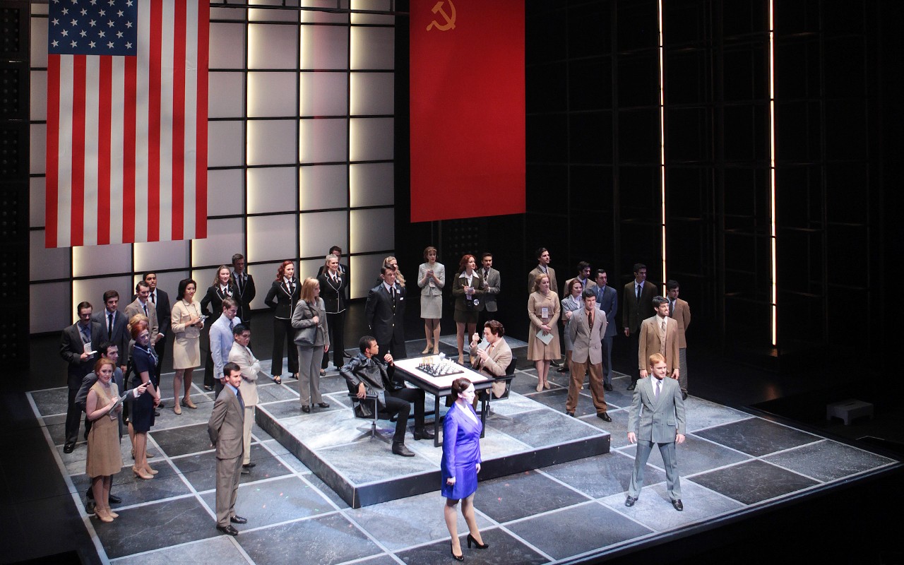 Student performers sing during a performance of Chess with American and Soviet flags hanging over a chess board floor 