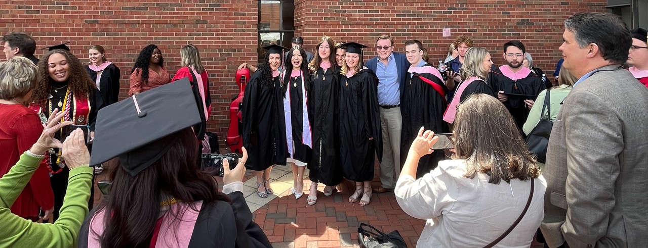 Students pose in cap and gown in front of the entrance to CCM's Corbett Center for the Performing Arts and Mary Emery Hall.